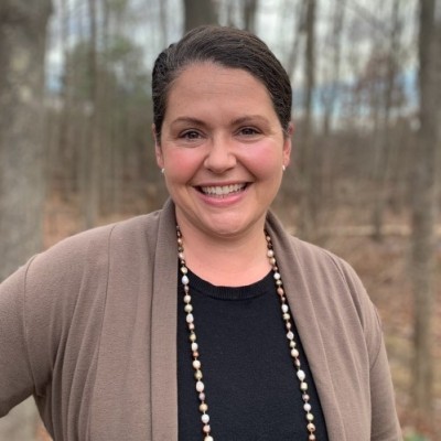 A white woman with short brown hair and wearing a black shirt and light brown sweater smiles