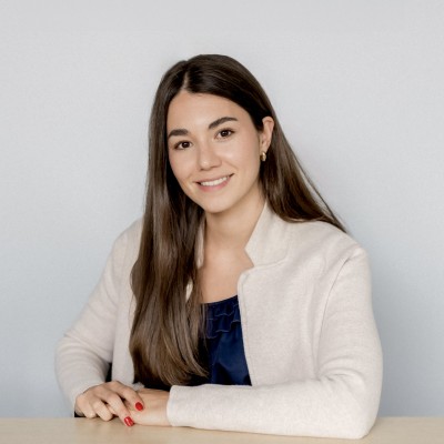 A smiling white woman with long brown hair and red fingernails sits at a wooden table in front of a white background