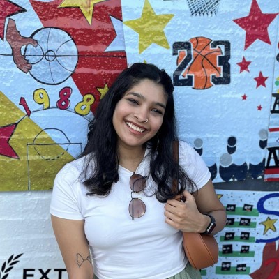 A smiling Indian woman with dark hair past her shoulders stands in front of a white brick wall painted with colorful graffiti