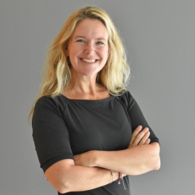 A smiling white woman with wavy blonde hair and wearing a black dress stands with her arms folded in front of a gray background