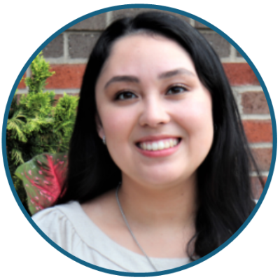 A woman with long, straight dark hair smiles while standing in front of a red brick wall with some greenery on it